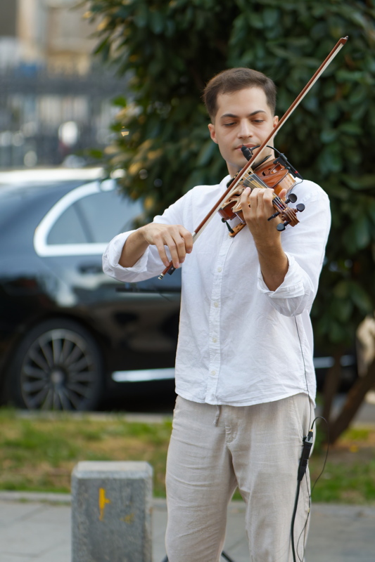 9 - A man wearing a white button-down shirt and light-colored trousers playing a violin outdoors near a vehicle and greenery